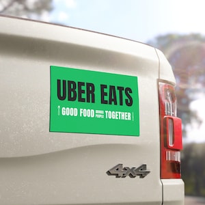 Peut inclure: Un aimant rectangulaire vert avec le texte "UBER EATS" en gras noir. En dessous, le texte blanc indique "GOOD FOOD BRINGS PEOPLE TOGETHER". L'aimant est fixé à l'arrière d'un véhicule blanc.