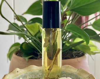 May include: A clear glass roller bottle with a black cap filled with a yellow liquid and dried herbs and flowers. The bottle is sitting on a light brown ceramic dish.