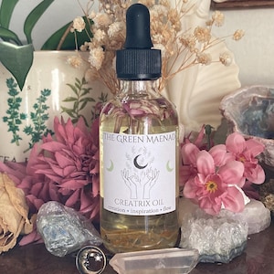 May include: A clear glass bottle with a black dropper top and a white label that reads "The Green Maenad Creatrix Oil creation - inspiration - flow". The bottle is filled with a light yellow liquid and has small pink flowers floating inside.