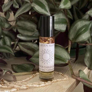May include: A small glass bottle with a black rollerball top. The bottle is filled with a clear liquid and contains small, dried flowers. The label on the bottle reads "Inner Child Oil" and "Green Maen". A sun symbol is also on the label.