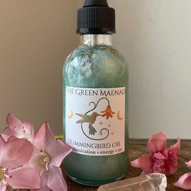May include: A clear glass bottle with a black dropper top, filled with shimmering green liquid. The label reads "THE GREEN MAENAD" and "HUMMINGBIRD OIL" with a hummingbird illustration. Pink flowers and a crystal are in the foreground.