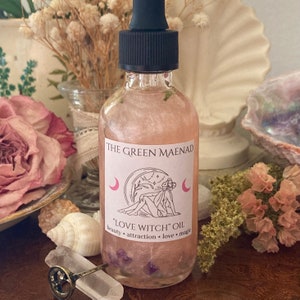 May include: A clear glass bottle with a black dropper top and a pink label that reads "The Green Maenad" and "Love Witch Oil" with the words "beauty, attraction, love, magic" underneath. The bottle is filled with a pink liquid and has small purple flowers inside.
