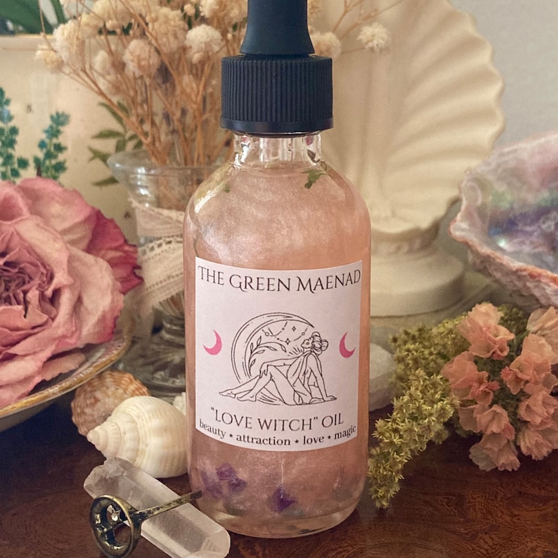 May include: A clear glass bottle with a black dropper top and a pink label that reads "The Green Maenad" and "Love Witch Oil" with the words "beauty, attraction, love, magic" underneath. The bottle is filled with a pink liquid and has small purple flowers inside.