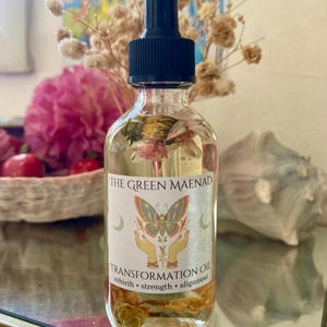 May include: A clear glass bottle with a black dropper top, filled with a golden liquid and dried flowers. The label reads "THE GREEN MAENAD TRANSFORMATION OIL" with a butterfly and hand graphic. The bottle is surrounded by dried flowers and a pink flower.