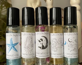 May include: Five clear glass roller bottles with black caps, each featuring a white label with the text Green Maena. The labels display illustrations of a starfish, a person, a moon, a hand with a coin, and hands with stars, labeled Mermaid Oil, Empress Oil, Lunar Witch Oil, Abundance, and Creativity.