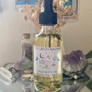 May include: A clear glass bottle with a black dropper top, filled with yellow liquid and botanical elements. The label reads "The Green Maenad Creativity Oil" with moon and hand illustrations. The bottle is surrounded by crystals and dried flowers.