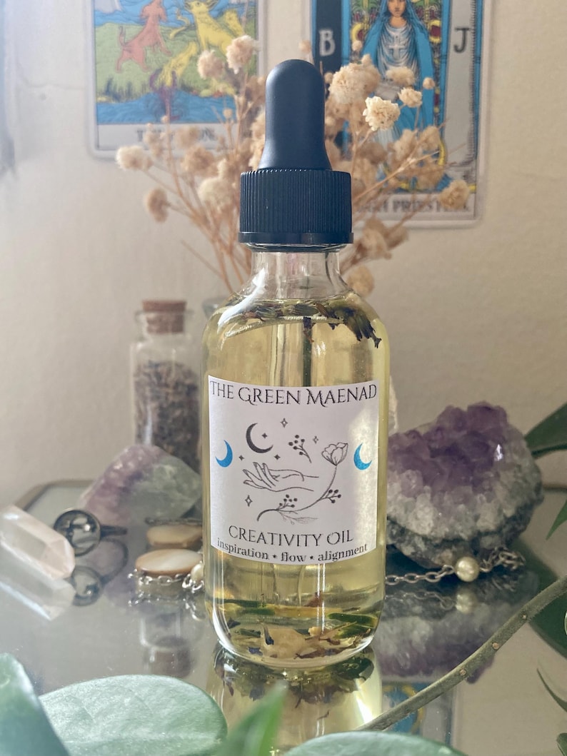 May include: A clear glass bottle with a black dropper top, filled with yellow liquid and botanical elements. The label reads "The Green Maenad Creativity Oil" with moon and hand illustrations. The bottle is surrounded by crystals and dried flowers.