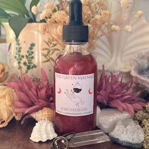 May include: A small, clear glass bottle with a red liquid inside. The bottle has a black dropper top and a white label that reads "The Green Maenad Sorceress Oil power - embodiment - creation".
