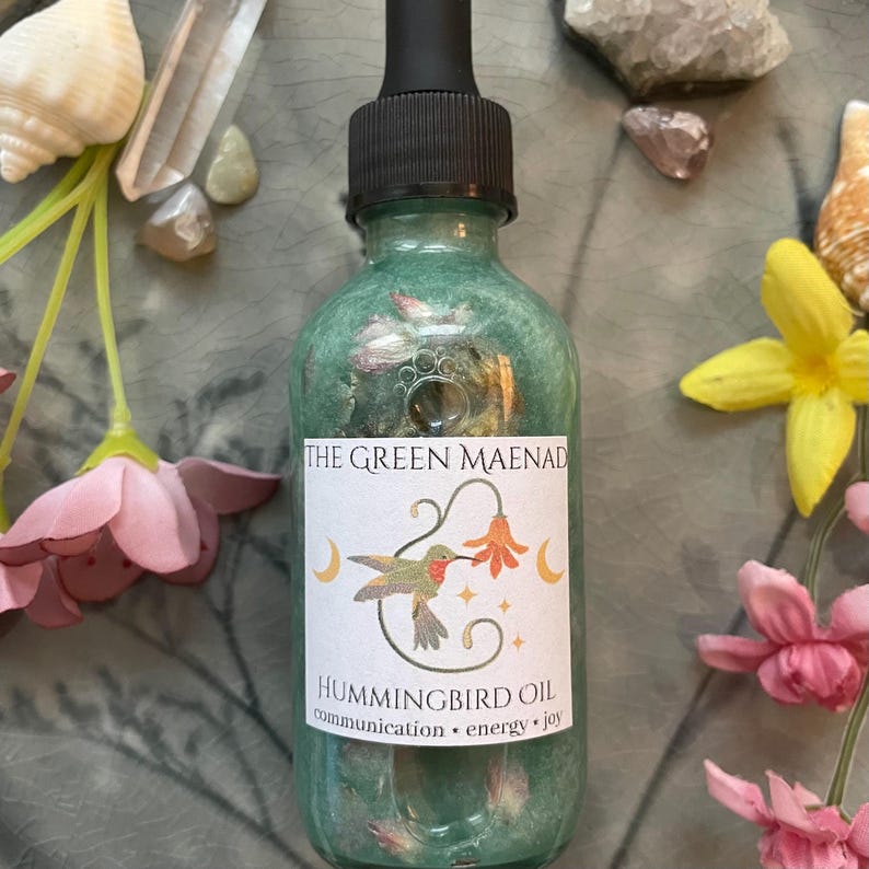 May include: A green glass bottle of Hummingbird Oil with a black dropper. The label reads "THE GREEN MAENAD" and features a hummingbird illustration. The bottle is surrounded by pink and yellow flowers, crystals, and seashells.