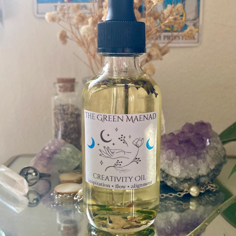 May include: A clear glass bottle of "Creativity Oil" with a black dropper top. The label reads "The Green Maenad" with a crescent moon and hand design. The oil is yellow with visible herbs. The bottle is surrounded by crystals and dried flowers.
