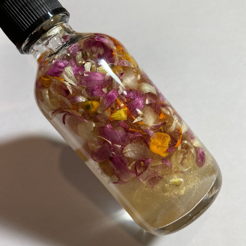 May include: A clear glass bottle filled with a golden liquid and various flower petals, including purple, white, and orange petals. The bottle has a black cap.