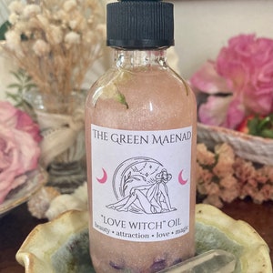 May include: A clear glass bottle with a black dropper top and a pink label that reads "THE GREEN MAENAD" and "LOVE WITCH" OIL. The label also includes a crescent moon and a drawing of a woman with a flower crown.