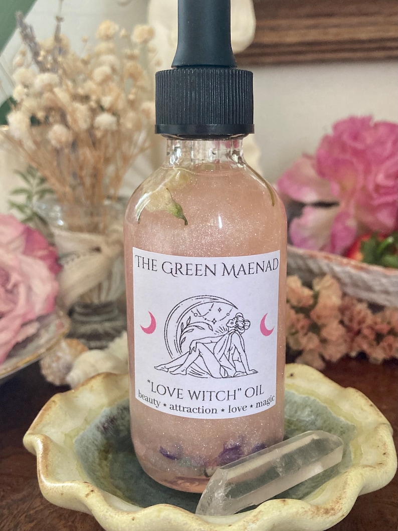 May include: A clear glass bottle with a black dropper top and a pink label that reads "THE GREEN MAENAD" and "LOVE WITCH" OIL. The label also includes a crescent moon and a drawing of a woman with a flower crown.