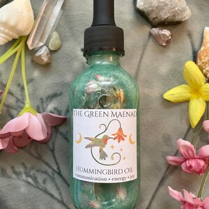 May include: A green glass bottle of Hummingbird Oil with a black dropper, labeled "The Green Maenad." The label features a hummingbird illustration and the words "communication, energy, joy." The bottle is surrounded by flowers, crystals, and seashells.