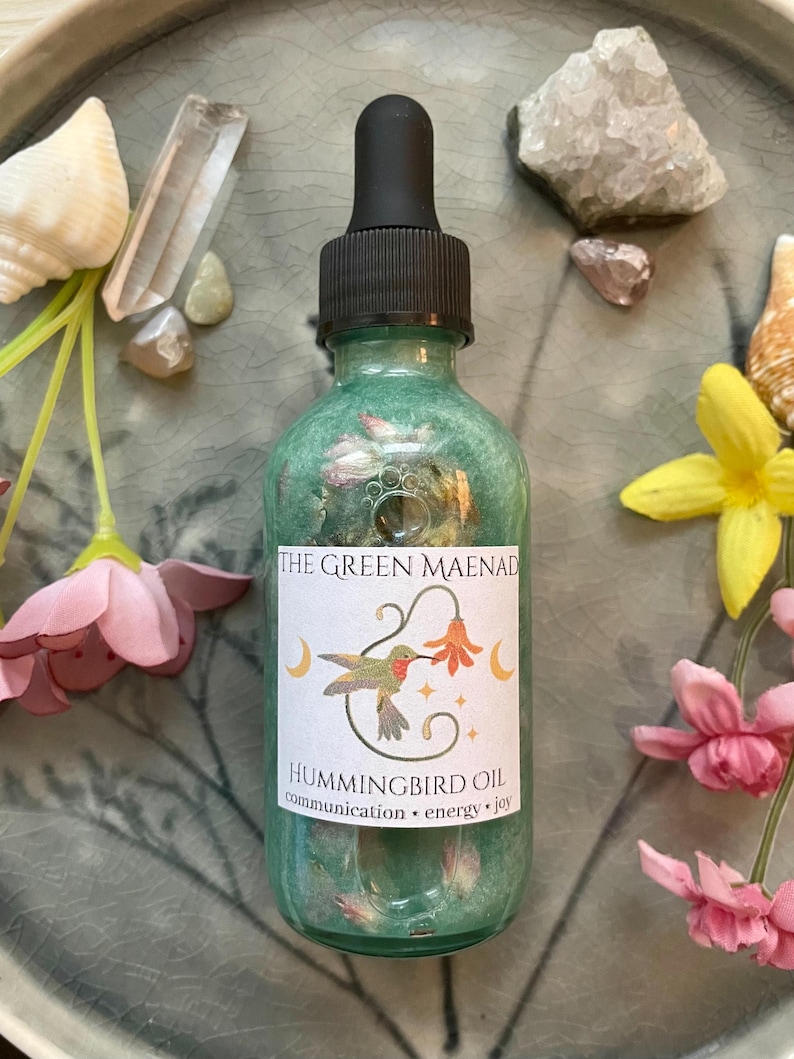 May include: A green glass bottle of Hummingbird Oil with a black dropper, labeled "The Green Maenad." The label features a hummingbird illustration and the words "communication, energy, joy." The bottle is surrounded by flowers, crystals, and seashells.