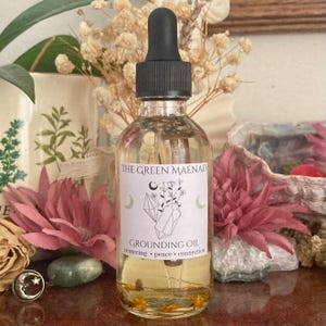 May include: A clear glass bottle with a black dropper top and a white label that reads "The Green Maenad Grounding Oil centering peace connection". The bottle is filled with a light yellow liquid and has small, yellow, flower-like objects floating inside.