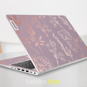 Könnte beinhalten: Ein Lenovo-Laptop mit einem floralen Lavendel-Design. Die Abdeckung des Laptops zeigt ein sich wiederholendes Muster goldfarbener botanischer Illustrationen. Die Tastatur ist schwarz und der Laptop ist auf einer weißen Oberfläche geöffnet. Das Lenovo-Logo ist sichtbar.