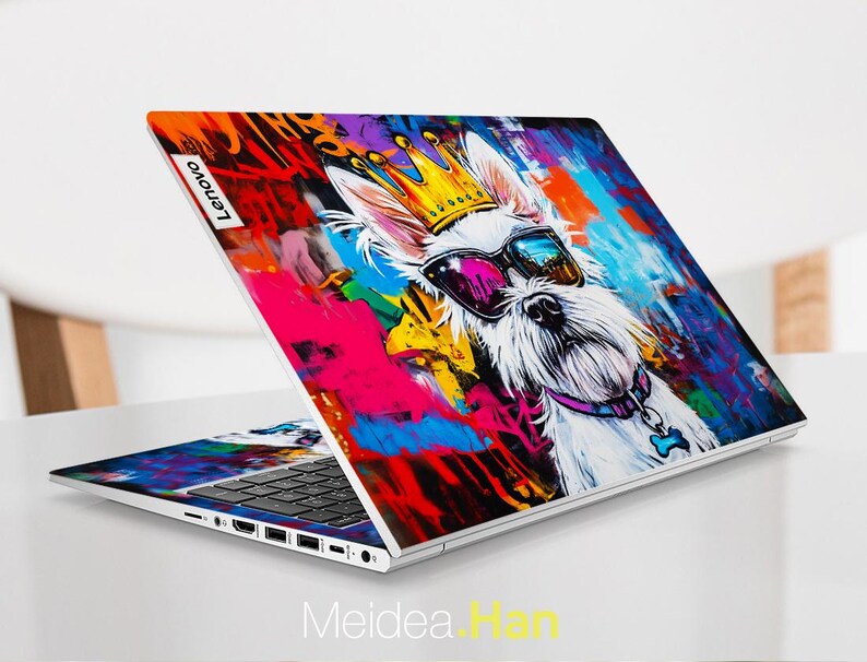 May include: A white laptop with a colourful, abstract design featuring a dog wearing sunglasses and a crown. The design includes splashes of pink, blue, yellow, and green paint. The laptop has the word "Lenovo" on the back.