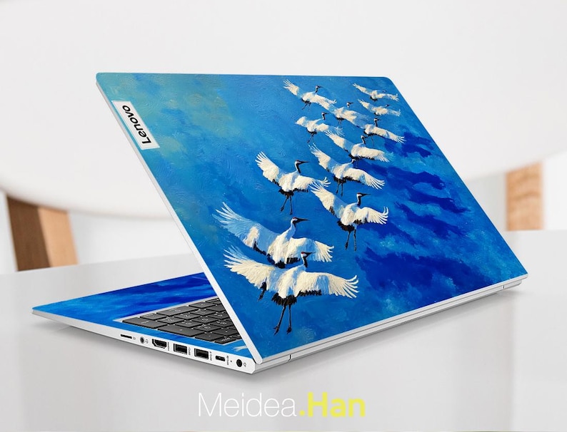 May include: A Lenovo laptop with a vibrant blue cover featuring a painting of white cranes in flight against a blue sky. The laptop is open, displaying the keyboard and screen. The Lenovo logo is visible.