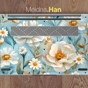 Puede incluir: Una piel para port&aacute;til con un dise&ntilde;o floral con flores blancas y hojas verdes sobre un fondo azul claro. El texto "Meidea.Han" est&aacute; impreso en amarillo en la parte superior de la piel.