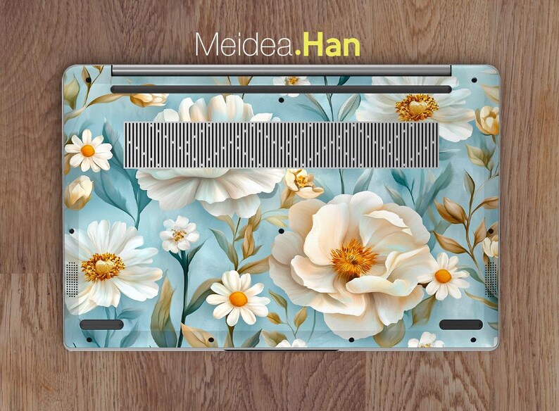 Puede incluir: Una piel para port&aacute;til con un dise&ntilde;o floral con flores blancas y hojas verdes sobre un fondo azul claro. El texto "Meidea.Han" est&aacute; impreso en amarillo en la parte superior de la piel.