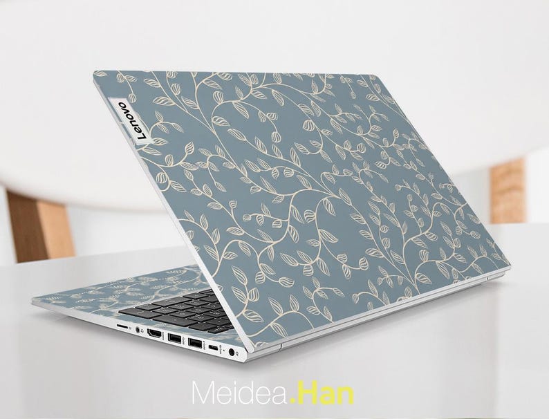 May include: A Lenovo laptop with a light blue cover featuring a repeating pattern of beige vines and leaves. The laptop is open, displaying the keyboard and ports. The Lenovo logo is visible on the top left corner.