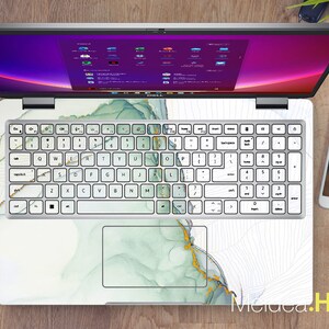 Może przedstawiać: Biały laptop z zielonym i złotym marmurowym wzorem na pokrywie i klawiaturze. Laptop jest otwarty, a ekran wyświetla pulpit z fioletowym tłem.