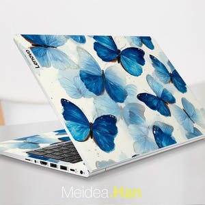 May include: A Lenovo laptop with a white cover featuring a pattern of blue butterflies. The butterflies vary in size and shade, creating a watercolor effect. The laptop is open, revealing the keyboard and screen.