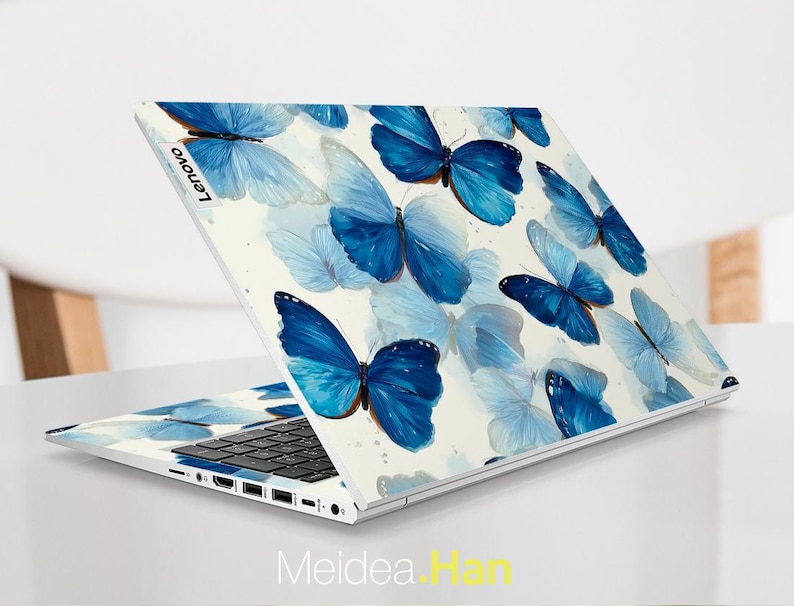 May include: A Lenovo laptop with a white cover featuring a pattern of blue butterflies. The butterflies vary in size and shade, creating a watercolor effect. The laptop is open, revealing the keyboard and screen.