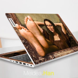 May include: A laptop computer with a custom skin featuring a painting of a woman in a brown dress with her feet up. The painting is framed in a gold frame. The laptop has a black keyboard and a silver body. The Lenovo logo is visible on the back of the laptop.