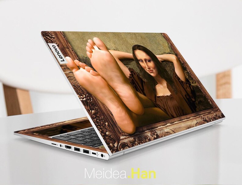 May include: A laptop computer with a custom skin featuring a painting of a woman in a brown dress with her feet up. The painting is framed in a gold frame. The laptop has a black keyboard and a silver body. The Lenovo logo is visible on the back of the laptop.