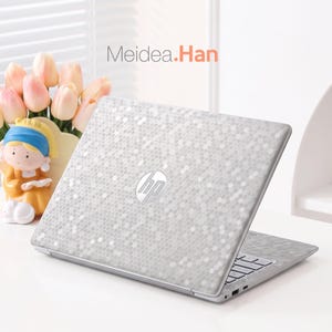Könnte beinhalten: Ein HP-Laptop mit einer silbernen Wabenmuster-Folie auf dem Deckel. Der Text Meidea.Han ist darüber zu sehen. Daneben steht eine kleine Figur mit blauem Kopftuch und gelber Kleidung, platziert neben einigen rosafarbenen Tulpenblüten.