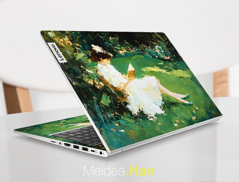 Puede incluir: Un port&aacute;til Lenovo con una cubierta personalizada que presenta una pintura impresionista de una persona reclinada en un campo verde, leyendo un libro. La obra de arte utiliza una paleta de verdes, blancos y amarillos. El port&aacute;til est&aacute; abierto, mostrando el teclado y la pantalla.