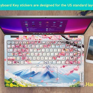 May include: A laptop computer with a pink and white cherry blossom design keyboard sticker. The sticker features a mountain scene with a blue sky and white clouds. The text "Lenovo" and "Meidea.Han" are visible on the laptop.