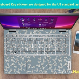 May include: A laptop with a floral keyboard skin. The keyboard keys are covered with stickers. The laptop is open, displaying a screen with a colorful interface. A smartphone and a watch are next to the laptop. The text "Keyboard Key stickers are designed for the US standard layout" is visible.