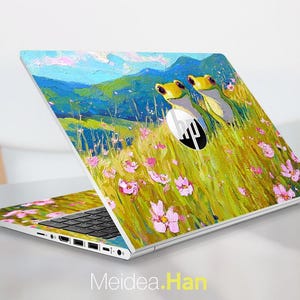 May include: A white HP laptop with a vibrant, impressionistic painting of two yellow-headed frogs in a field of pink flowers and green grass. The background features blue mountains and sky. The laptop's keyboard is visible.