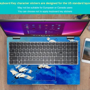 May include: Laptop with a keyboard skin featuring a blue and white crane design. The keyboard has standard US layout keys. The screen displays a colorful interface. A smartphone and watch are visible on the wooden surface.