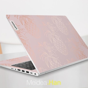 Puede incluir: Un portátil Lenovo con una cubierta rosa claro con un patrón repetido de piñas doradas. El portátil está abierto, mostrando el teclado y la pantalla. El logotipo de Lenovo es visible en la esquina superior izquierda.