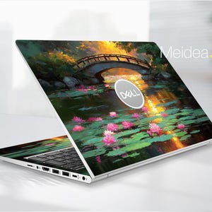 May include: A Dell laptop with a custom skin featuring a serene landscape painting. The artwork depicts a bridge over a pond with lily pads and pink flowers, bathed in warm sunlight. The laptop is open, displaying the keyboard and ports.