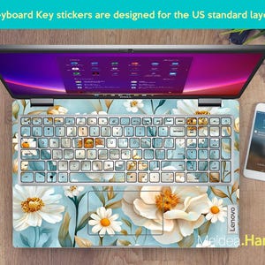 Puede incluir: Un ordenador port&aacute;til con una pegatina de teclado floral. La pegatina presenta flores blancas y amarillas sobre un fondo azul. La pantalla del port&aacute;til muestra un escritorio con iconos. El texto "Keyboard Key stickers are designed for the US standard layout" es visible en la parte superior de la imagen.