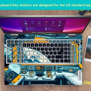 May include: A laptop computer with a keyboard sticker featuring a space-themed design. The sticker has a blue, yellow, and black color scheme and depicts a spaceship cockpit.
