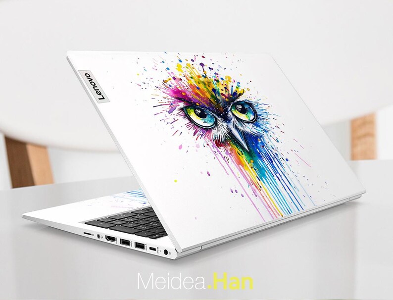Pu&ograve; includere: Laptop bianco con un colorato design a gufo ad acquerello. L'opera d'arte presenta gli occhi di un gufo e uno spruzzo di colori arcobaleno. Il laptop ha una tastiera nera e il logo Lenovo.