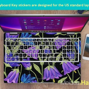 May include: Laptop with keyboard key stickers designed for the US standard layout. The keyboard has a floral design with purple and blue bellflowers on a black background. The laptop is open, displaying a colorful screen.
