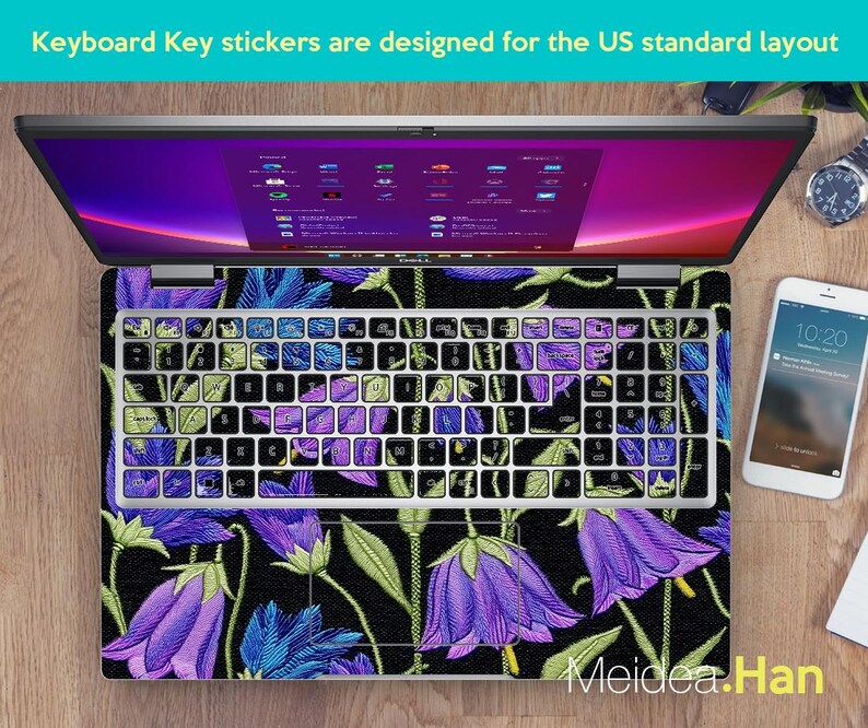 May include: Laptop with keyboard key stickers designed for the US standard layout. The keyboard has a floral design with purple and blue bellflowers on a black background. The laptop is open, displaying a colorful screen.