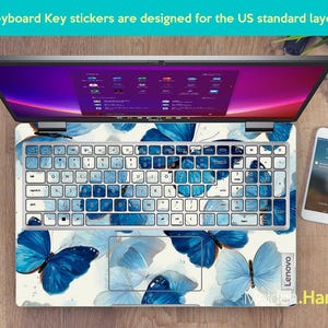 May include: Laptop with a keyboard overlay featuring blue butterfly designs. The keyboard keys are white with blue accents. The laptop screen displays a purple and pink interface. A smartphone and watch are visible.