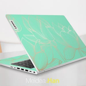 May include: A silver laptop with a mint green and gold floral design on the lid. The design features delicate, stylized lotus flowers. The laptop has a black keyboard and a silver trackpad. The Lenovo logo is visible on the lid.