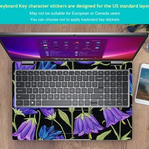May include: Laptop with a floral keyboard skin featuring purple bellflowers and green leaves on a black background. The keyboard has a standard US layout. A smartphone and watch are visible on the wooden surface.