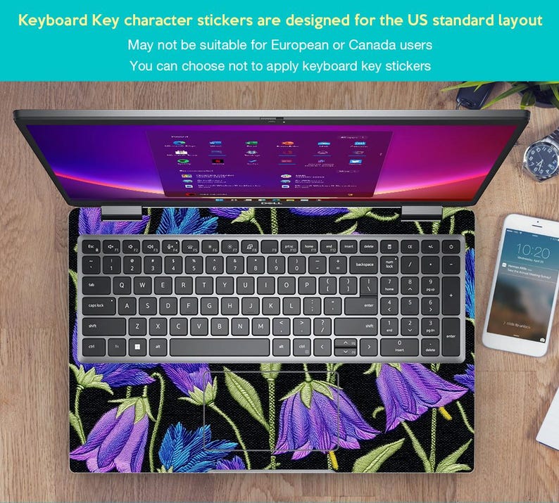 May include: Laptop with a floral keyboard skin featuring purple bellflowers and green leaves on a black background. The keyboard has a standard US layout. A smartphone and watch are visible on the wooden surface.