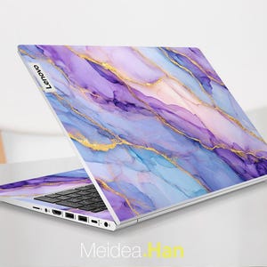 Puede incluir: Un portátil Lenovo con un diseño abstracto en tonos morados, azules y dorados. La superficie del portátil tiene un efecto marmolado. El logotipo de Lenovo es visible. El portátil está abierto, mostrando el teclado.