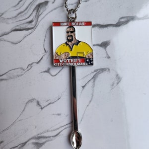 May include: A silver chain necklace with a silver spoon pendant. The spoon has a rounded bowl and a long handle. The pendant is attached to a square metal charm with a cartoon image of a man wearing a yellow shirt and sunglasses. The charm has the text "MIKE NOLAN VOTE #1 CITY COUNCILMAN" on it.